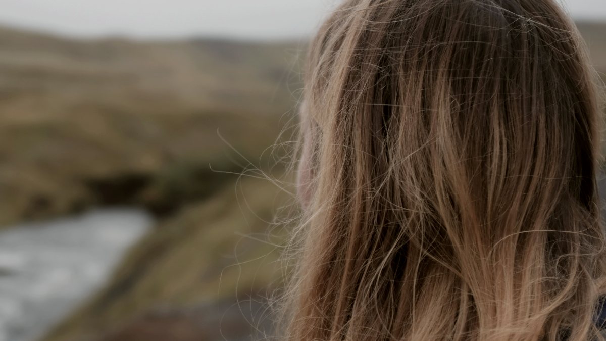 
a blonde woman facing a body of water at daytime with hills in the background by isuru perera
