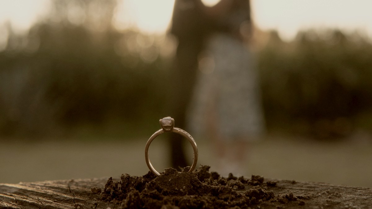 
A couple standing in front of each other in the background with a wedding ring standing in dirt in focus by Silvestre Leon
