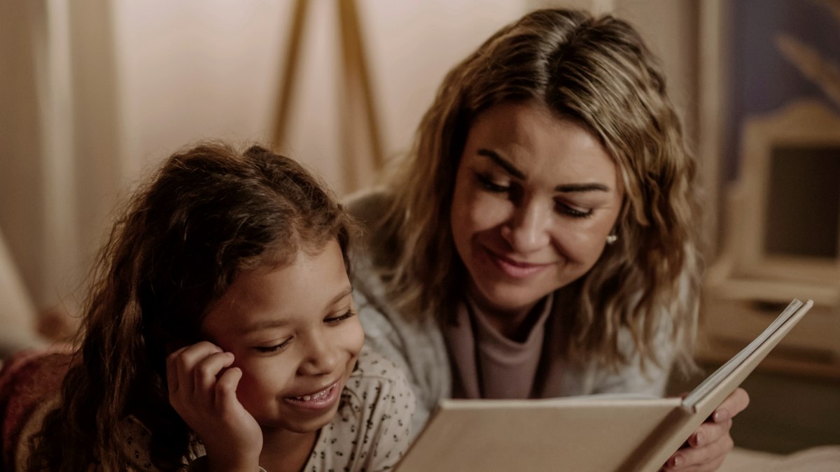 
A happy mother with her little daughter lying on bed and reading book in evening at home
