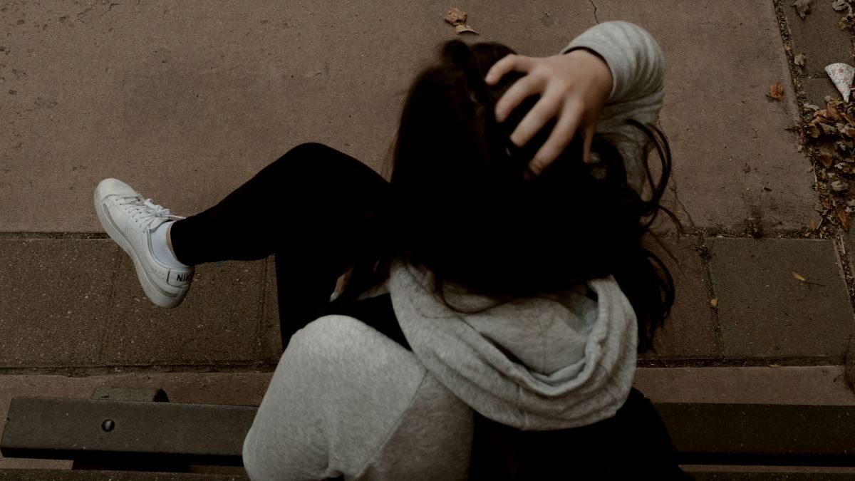 
a long haired woman sitting on a bench on a sidewalk wearing a backpack by Julia Rodriguez
