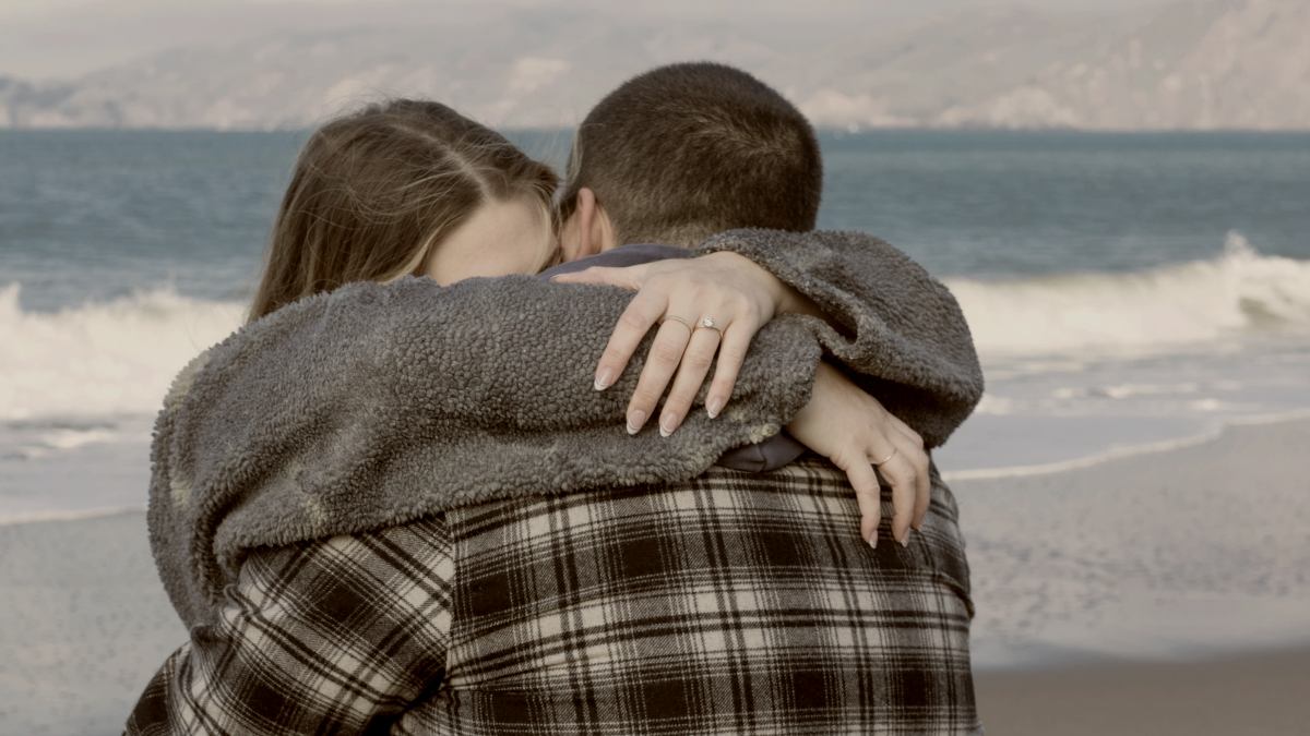 
a man and a woman hugging on the beach with mountain range in the background by Felicia Montenegro
