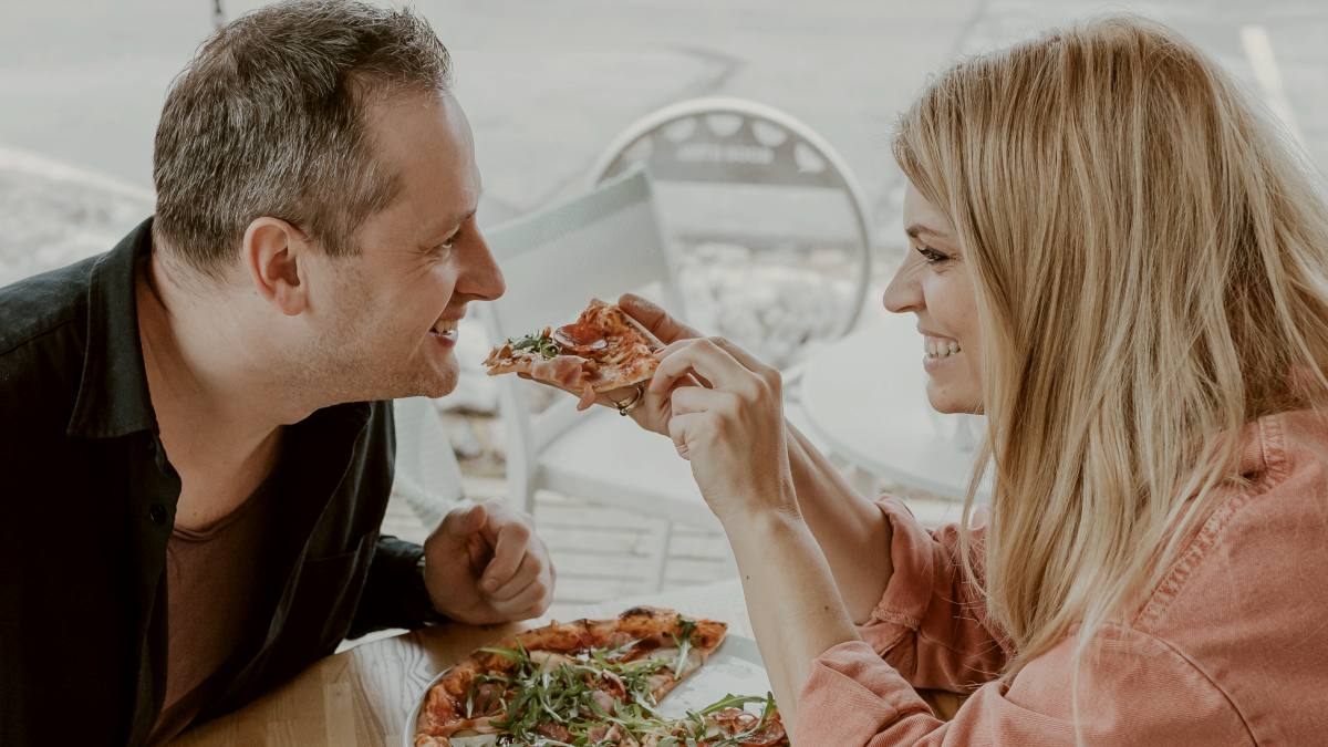 
a man and a woman sitting at a table eating pizza smiling by Daiga Ellaby
