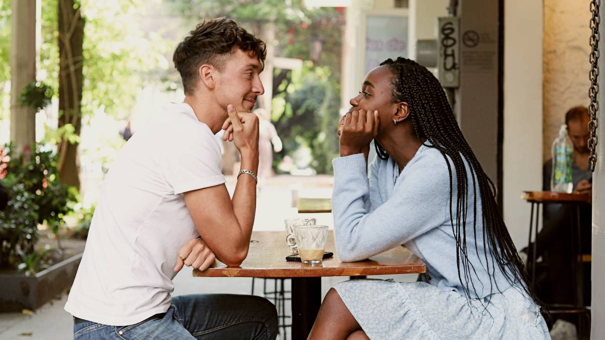 
a man and a woman sitting at a table outside on a date by good faces
