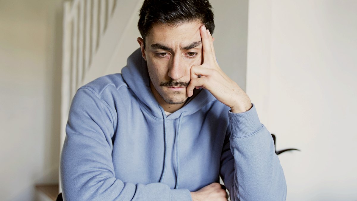 a man with light blue hoodie sitting at a table with his hand on his face by Guillaume Issaly