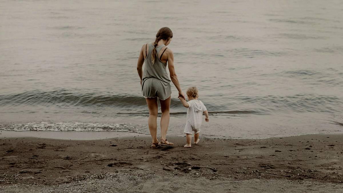 
a mother holding her toddler's hand as they walk on the sea shore
