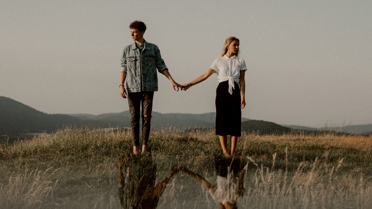 
A portrait of young couple standing in nature in countryside, holding hands but looking away from each other.
