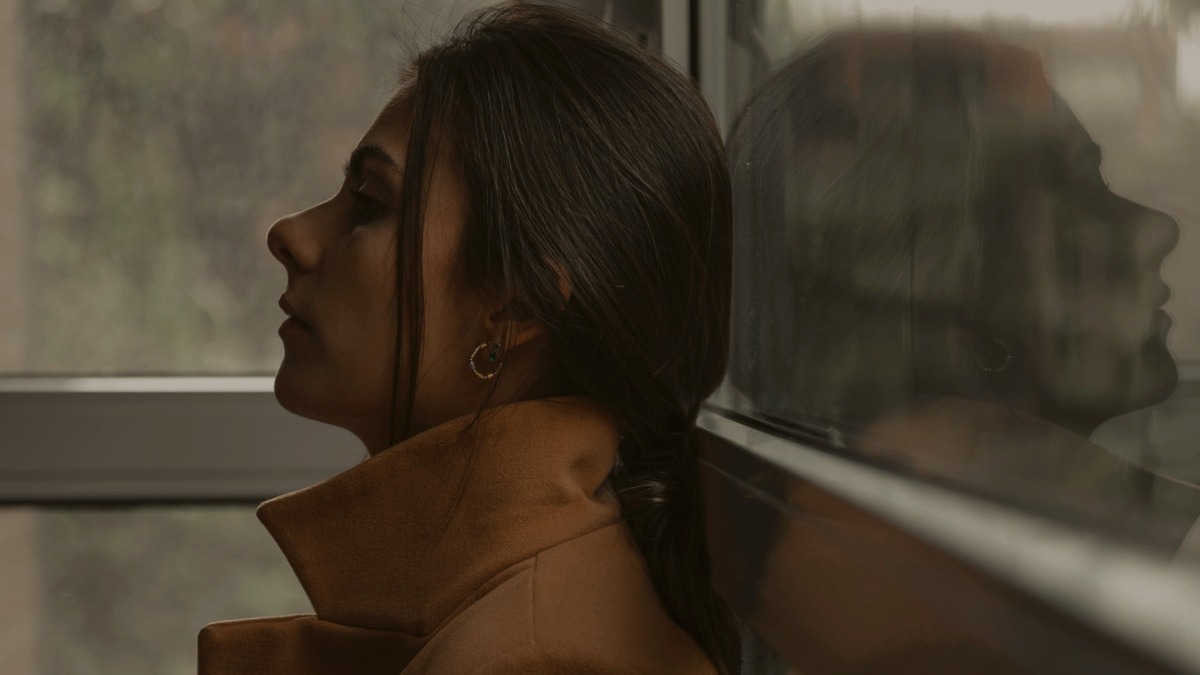 
a woman in a tan coat standing with her back to a window, her image is reflected back at her  by Pablo Merchán Montes
