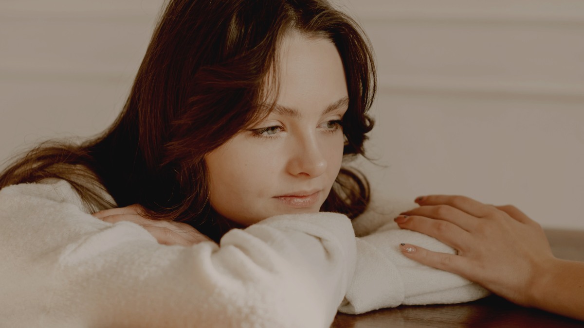 a woman sitting at a table in a white sweater and a comforting hand on her elbow by Kateryna Hliznitsova