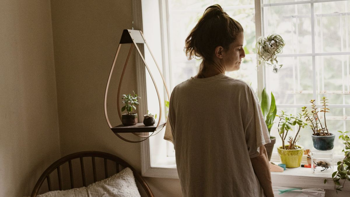 a woman standing in a white tshirt in front of a window looking out in a bedroom by Daniel Martinez