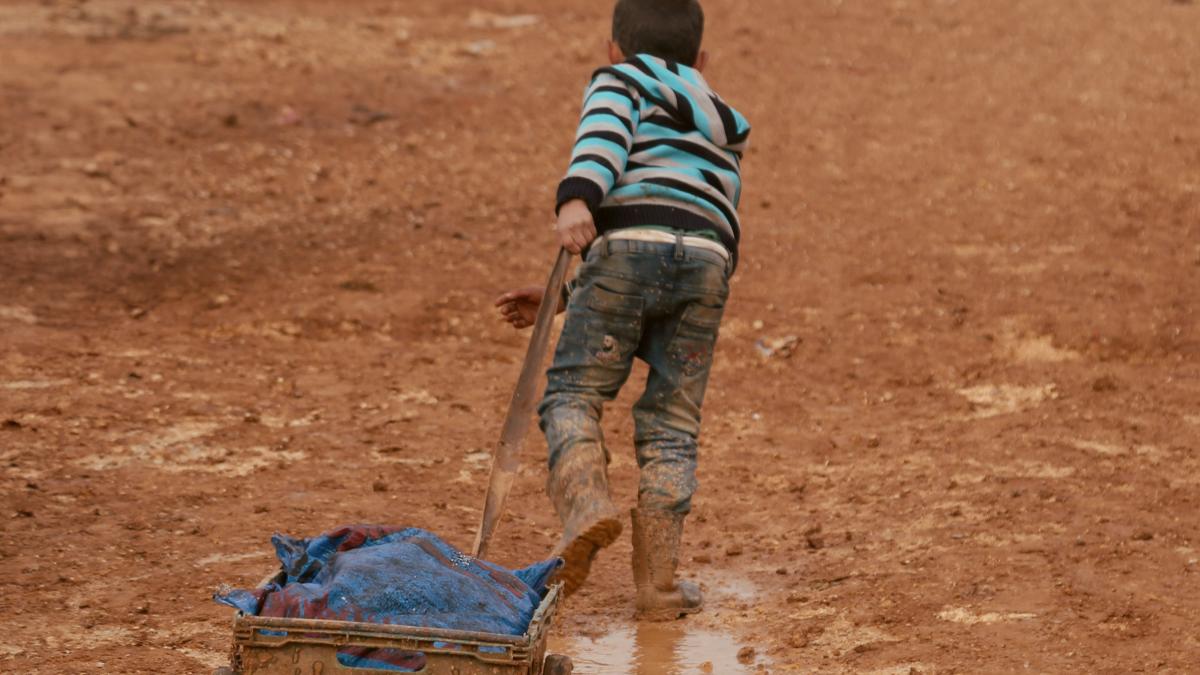 
A young boy in gum boots pulling a wagon with blue bags through a muddy dirt field by salah darwish
