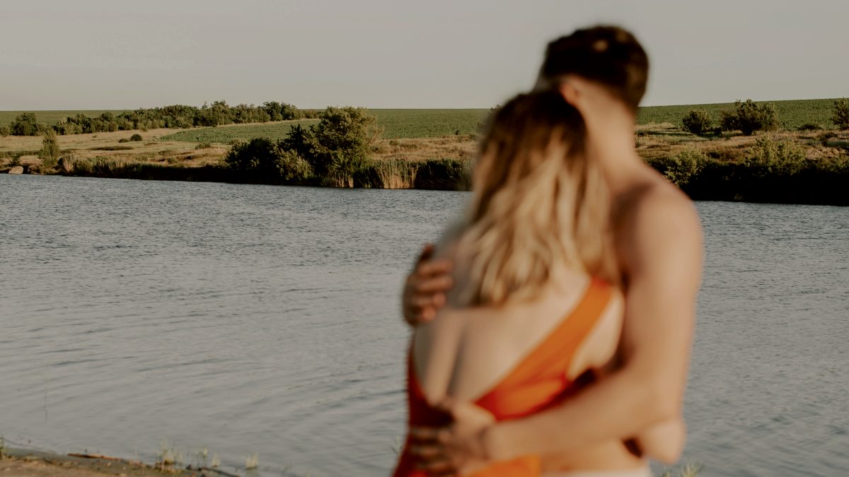 
an out of focus man and woman embracing on the shore of a lake by Kateryna Hliznitsova
