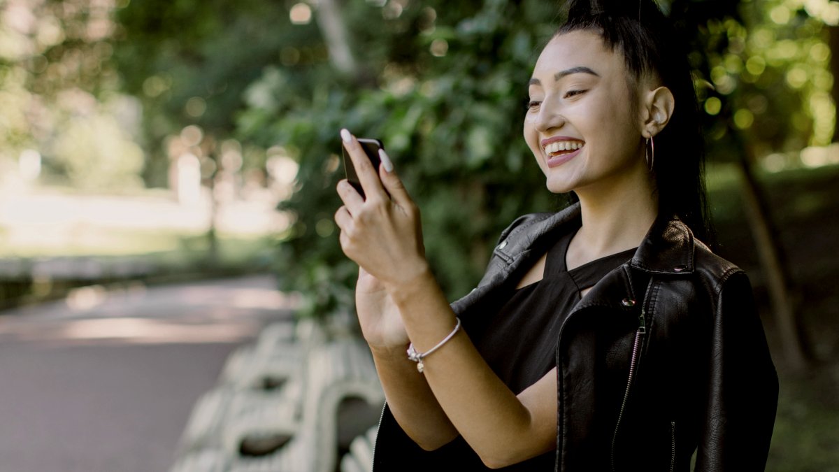 
Lovely cheerful young Asian brunette woman, wearing trendy black outfit, sitting on the bench at the city park, holding mobile phone, while reading funny memes or message.
