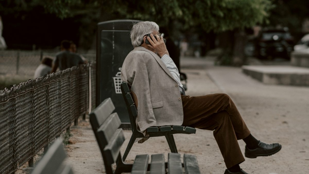 
man in brown jacket sitting on a green park bench with a cellphone pressed against his ear by Thomas de LUZE

