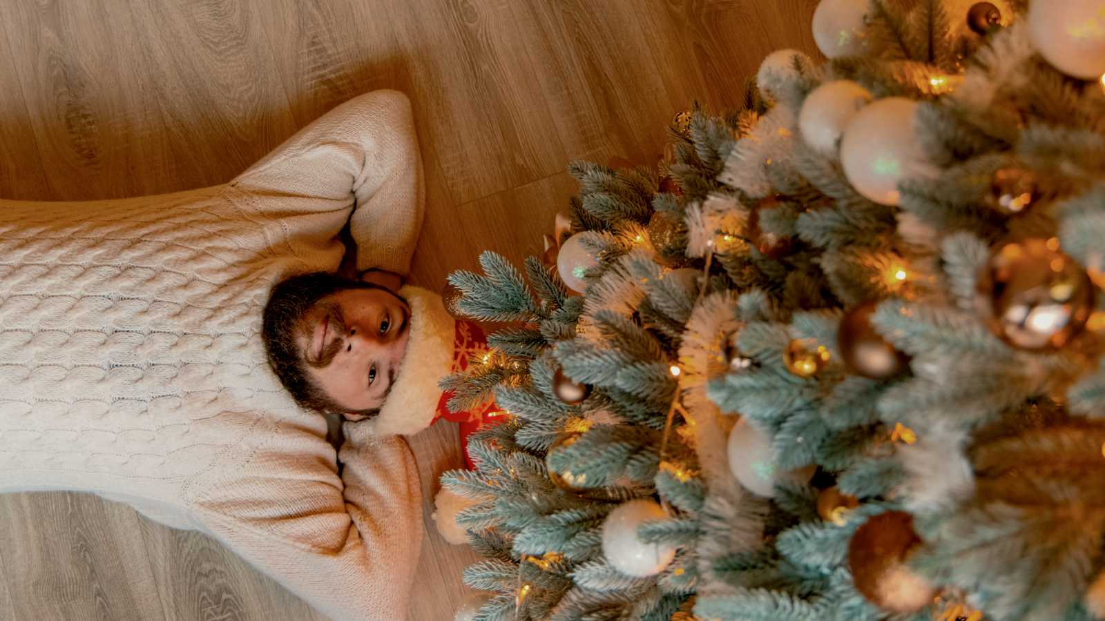 
man in white long sleeve shirt lying on floor under a christmas tree by Artem Kniaz
