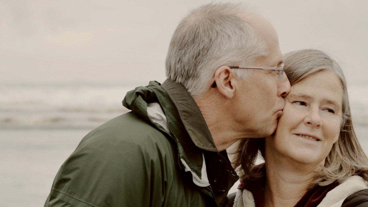
older man in green jacket kissing older woman in black jacket on cheek beside the ocean by esther ann
