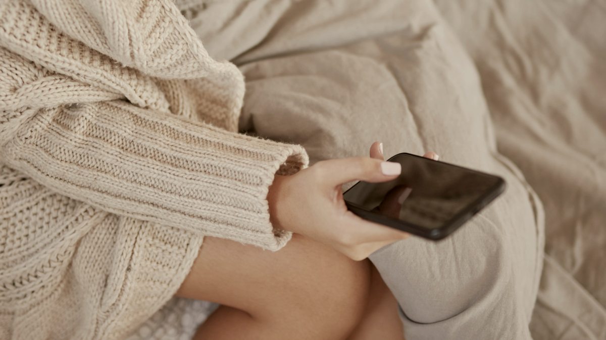 
Top view of a woman in beige sweater sitting on bed and using smart phone
