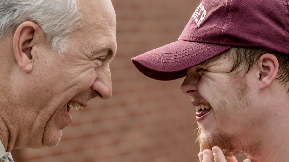 
two men laughing at each other, one wearing a baseball cap and another elderly by Nathan Anderson
