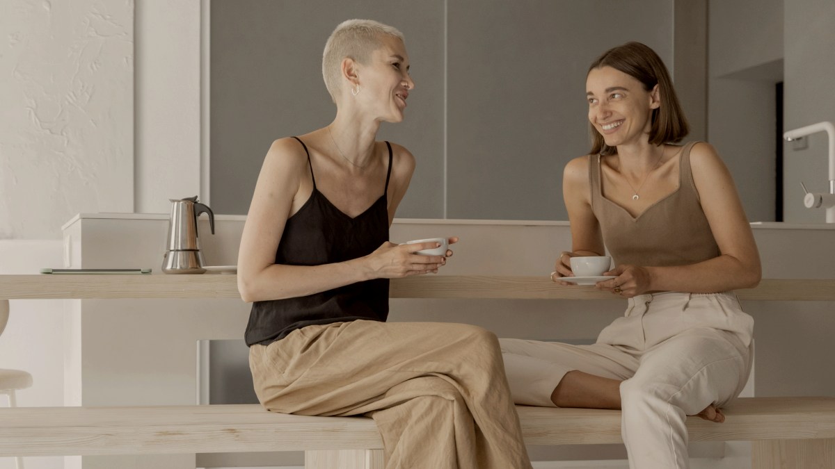 
two women drinking coffee, sitting together and talking at modern kitchen in studio apartment
