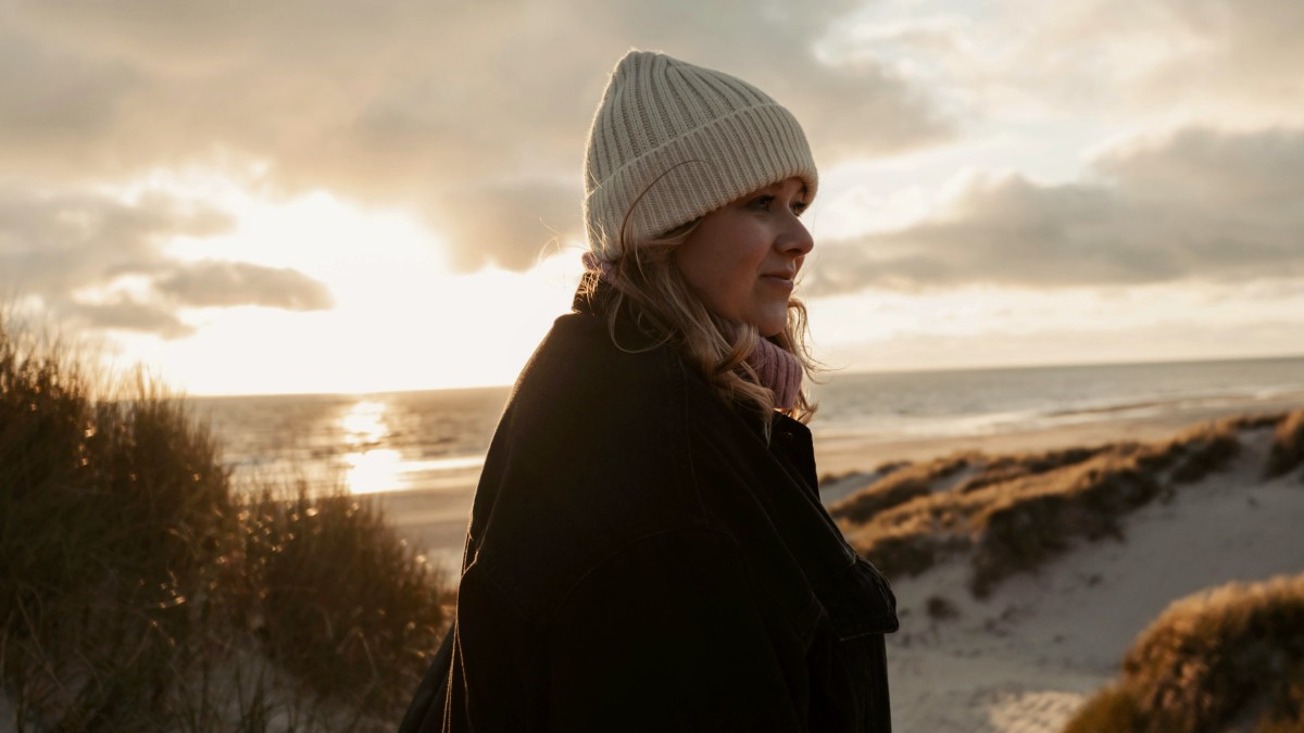 
Woman in beige beanie and black jacket enjoys a beach view during sunset by Lukas Zischke

