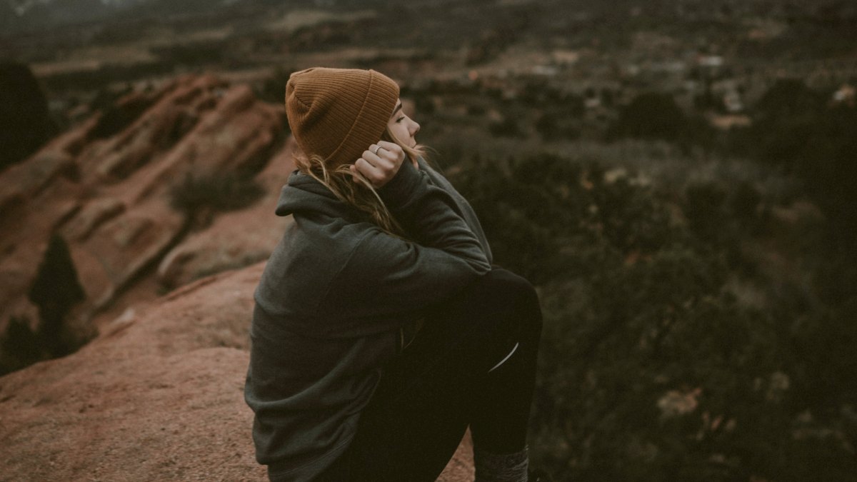 
woman in green jacket and a brown beanie sitting on a cliff
