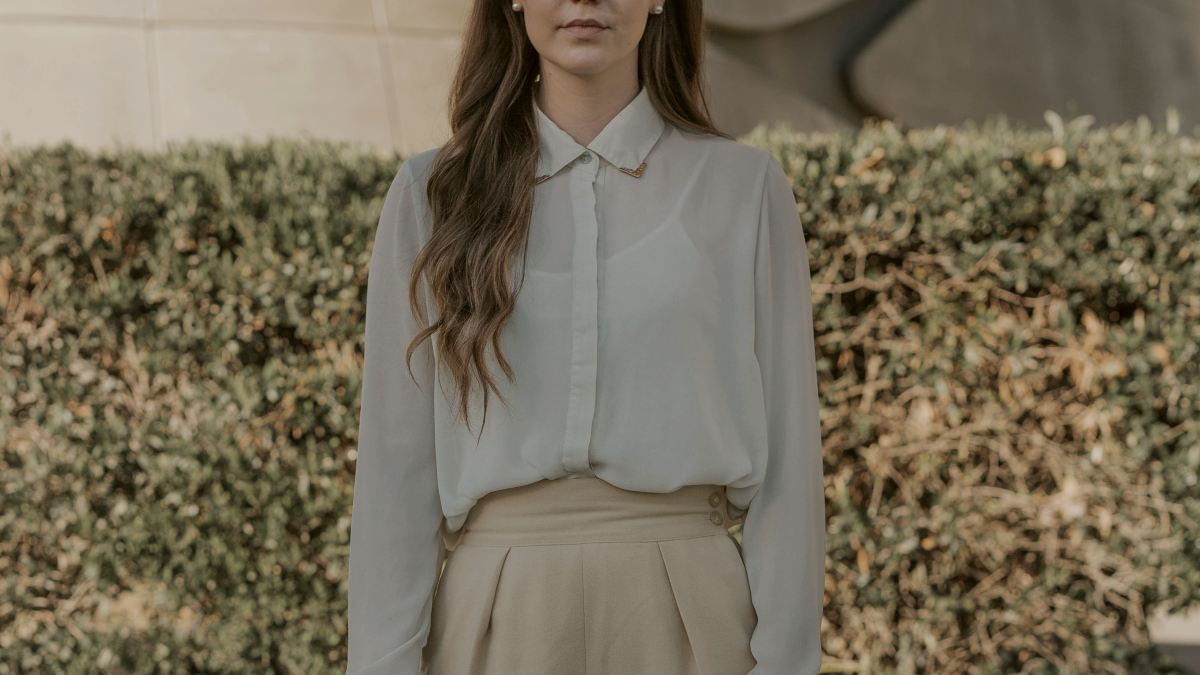 
woman wearing gray button-up long-sleeved shirt and brown pants standing in front of bushes
